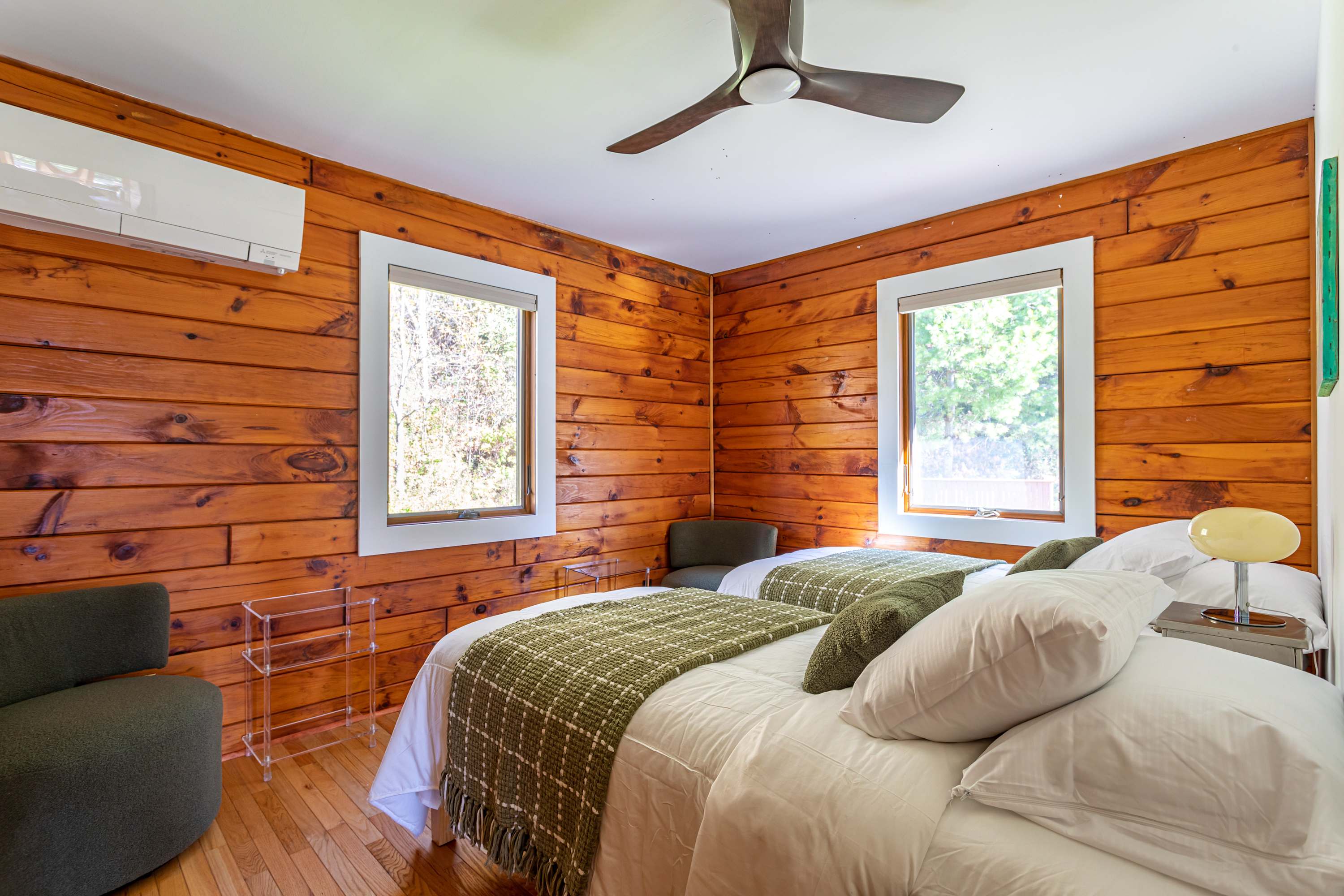 Another view of bedroom-2 showing two twin beds with green throws, a wood-paneled wall, and an air conditioning unit. Two windows provide natural light.