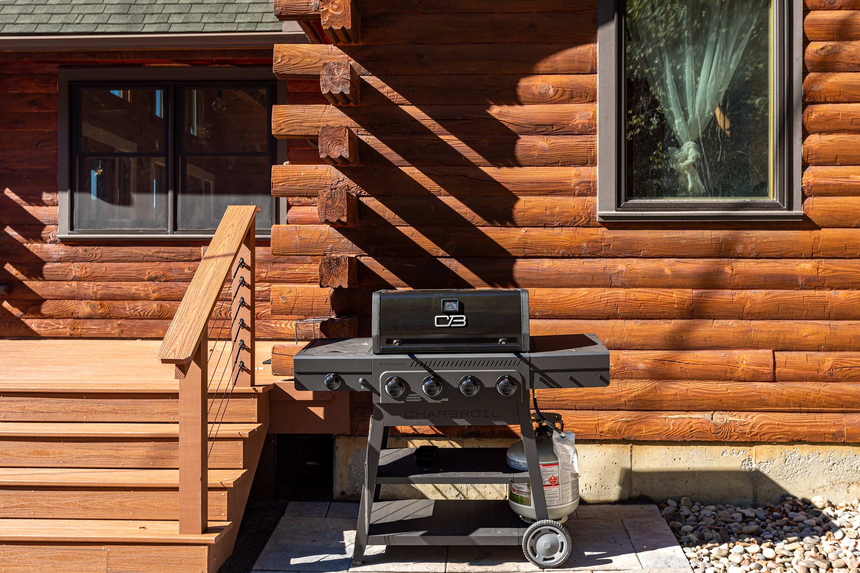Outdoor grilling area featuring a black Char-Broil gas grill with a propane tank, situated on a stone patio next to the log cabin exterior. Wooden stairs lead up to a deck.