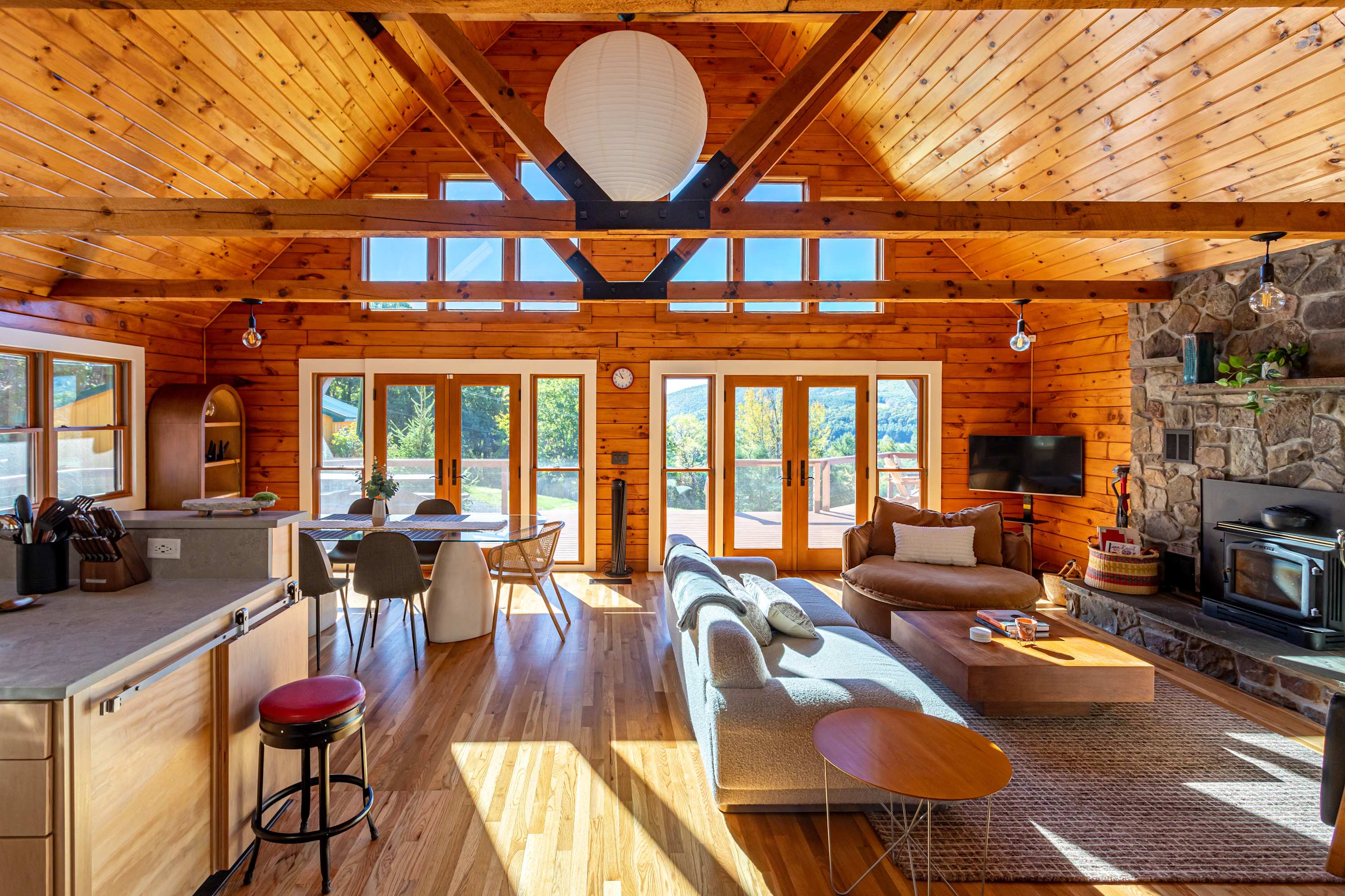 Open-concept living and dining area with vaulted wood-paneled ceilings and exposed beams, featuring a stone fireplace, comfortable seating, and large glass doors leading to a deck with mountain views.