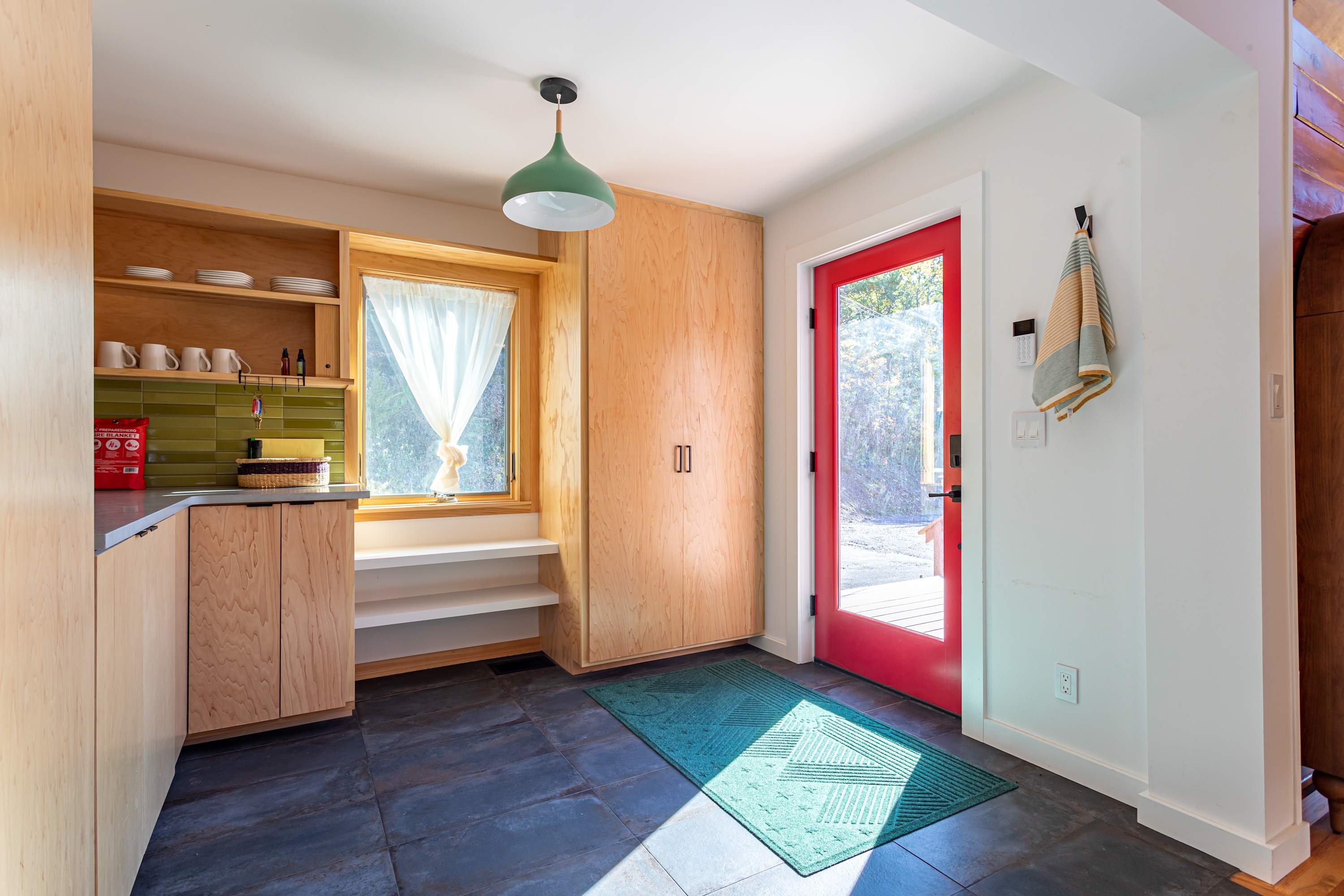 Bright entry area with a vibrant red door, dark slate floor tiles, light wood built-in cabinets and shelving, and a small window with white curtains, illuminated by a green pendant light.
