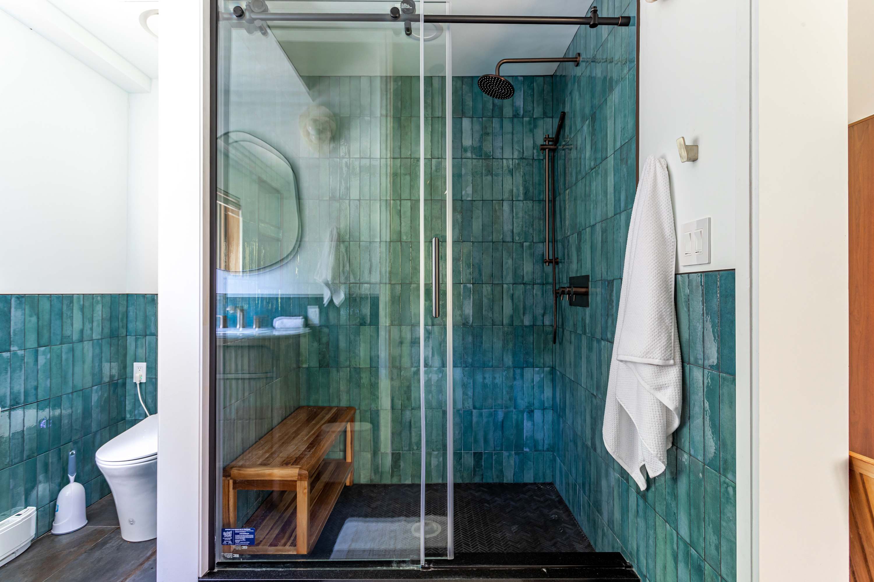 Close-up of bathroom-1's walk-in shower with teal subway tiles, a rainfall showerhead, and a wooden bench. A modern toilet and a small round mirror are also visible.