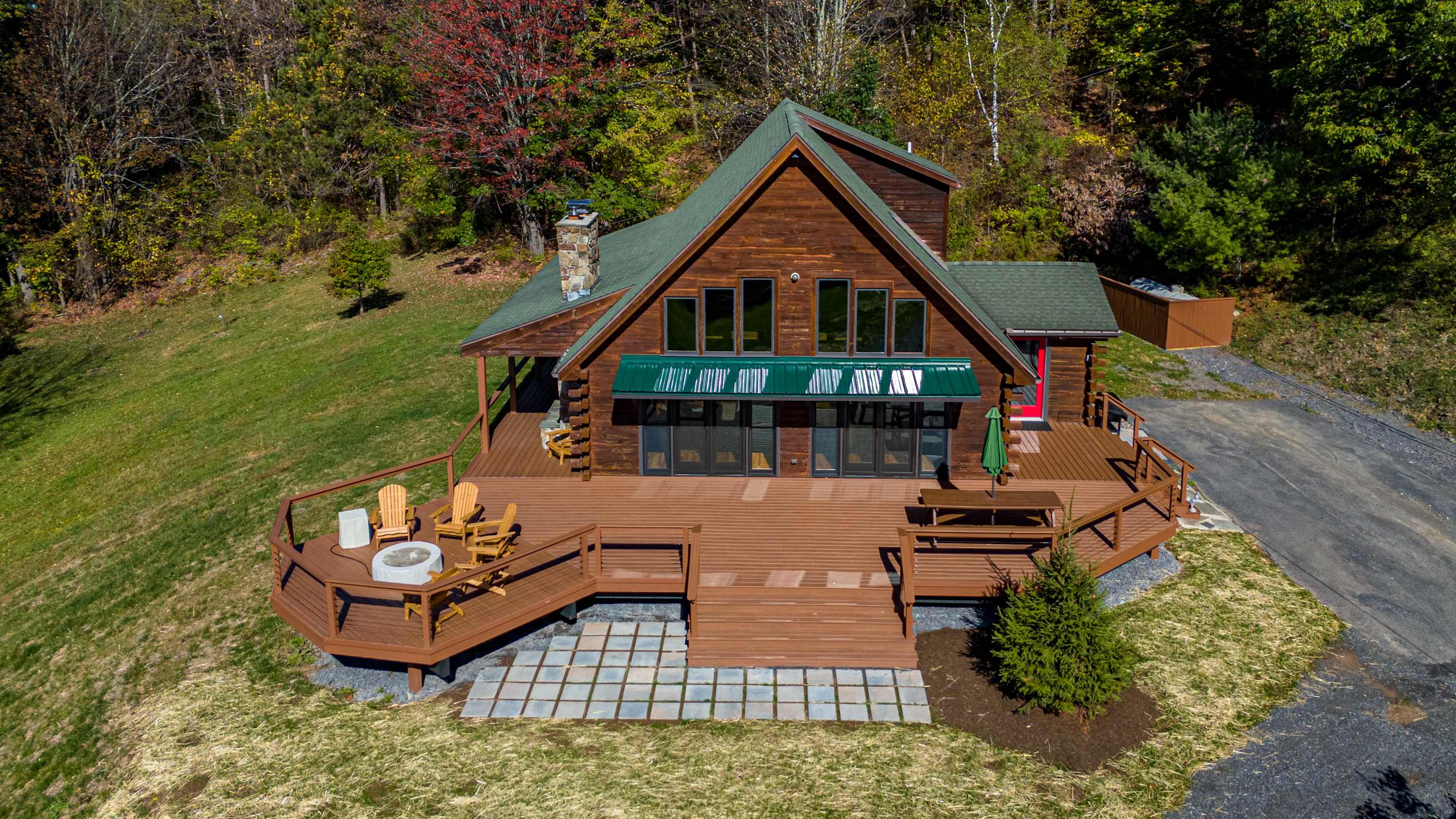 Aerial view of a rustic log cabin with a green roof and extensive wraparound wooden deck, surrounded by lush green trees and a gravel driveway. The deck features outdoor seating and a fire pit.