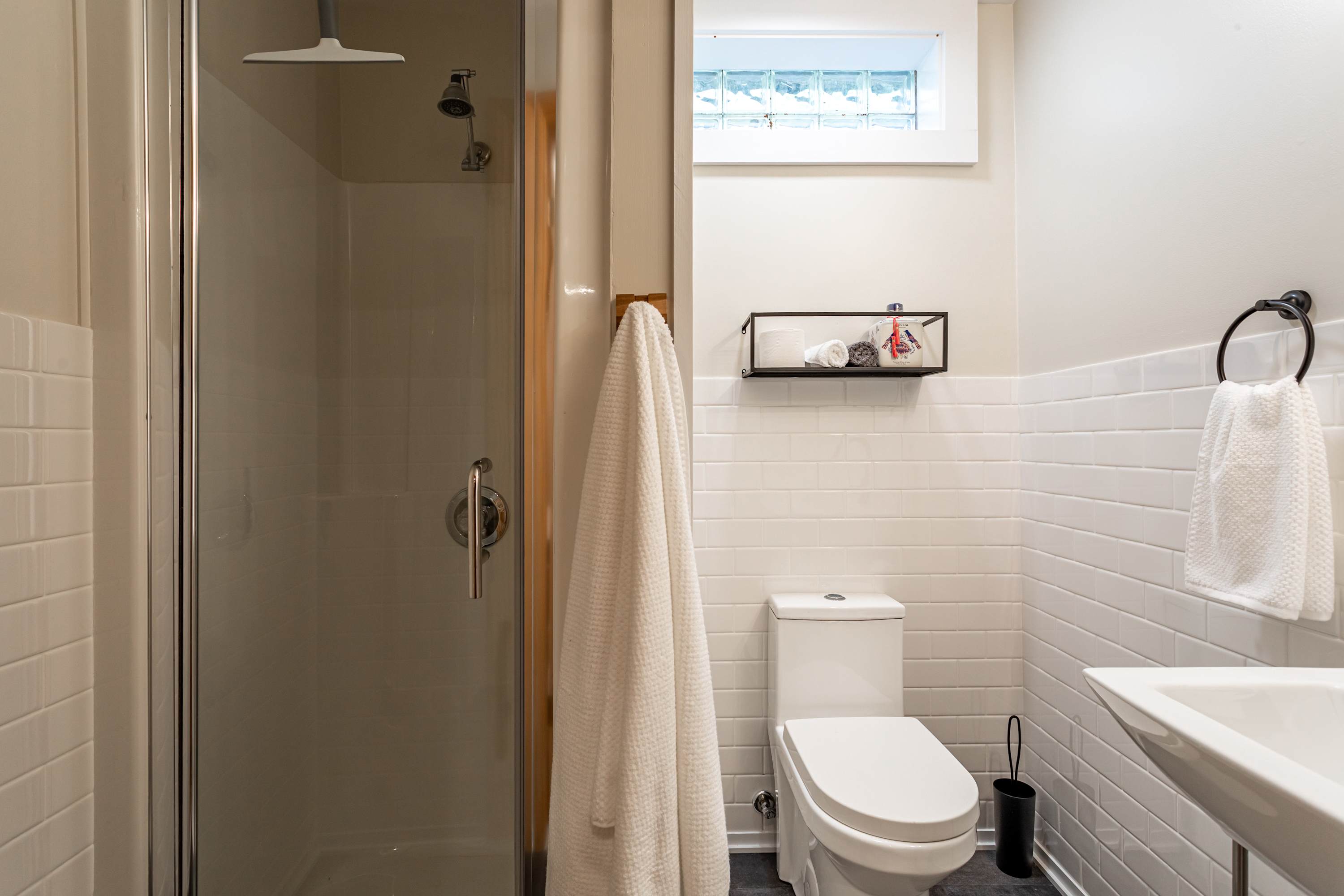 Modern bathroom featuring a glass-enclosed shower, white subway tile walls, a white toilet, and a wall-mounted sink. A small window with glass blocks provides natural light.