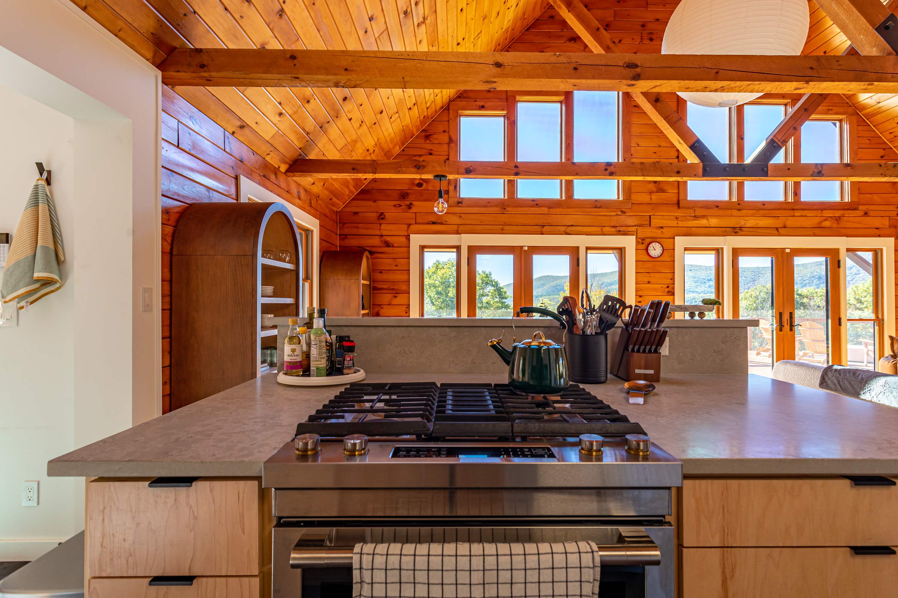 Kitchen island with a built-in stainless steel gas range and oven, light gray countertop, and light wood cabinetry, offering a view of the expansive living area and mountain landscape through large windows.