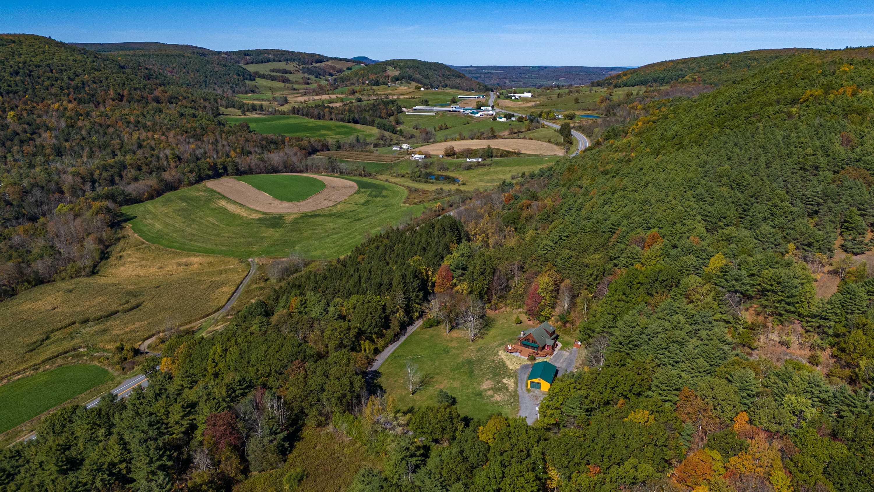 Aerial view showcasing the wooden cabin nestled among lush green and autumn-colored trees in a valley, surrounded by rolling hills, fields, and distant roads.