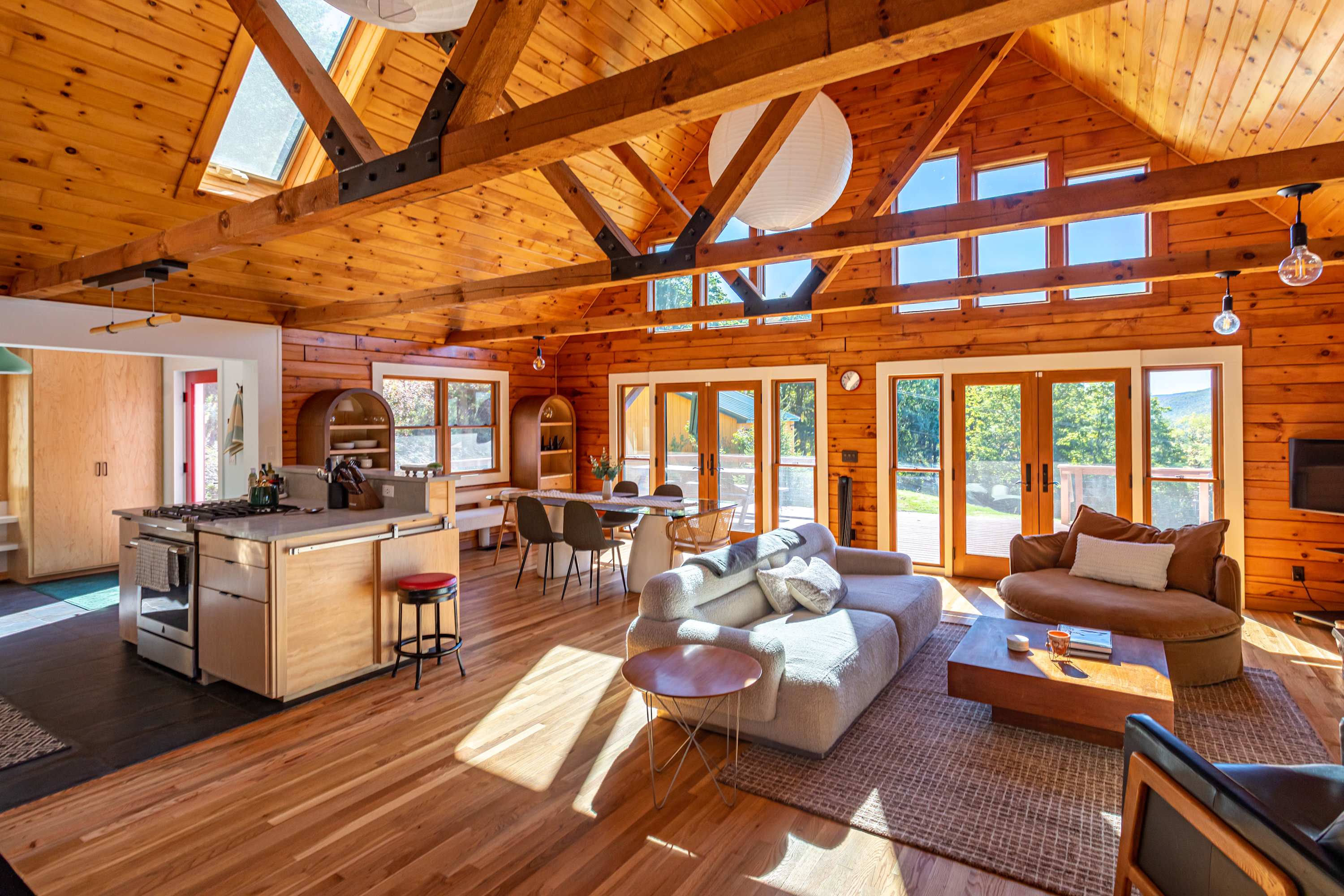 Expansive open-concept living space featuring a light gray sectional sofa, a wooden coffee table, and a dining area, all under a high vaulted ceiling with exposed wooden beams and large windows offering mountain views.