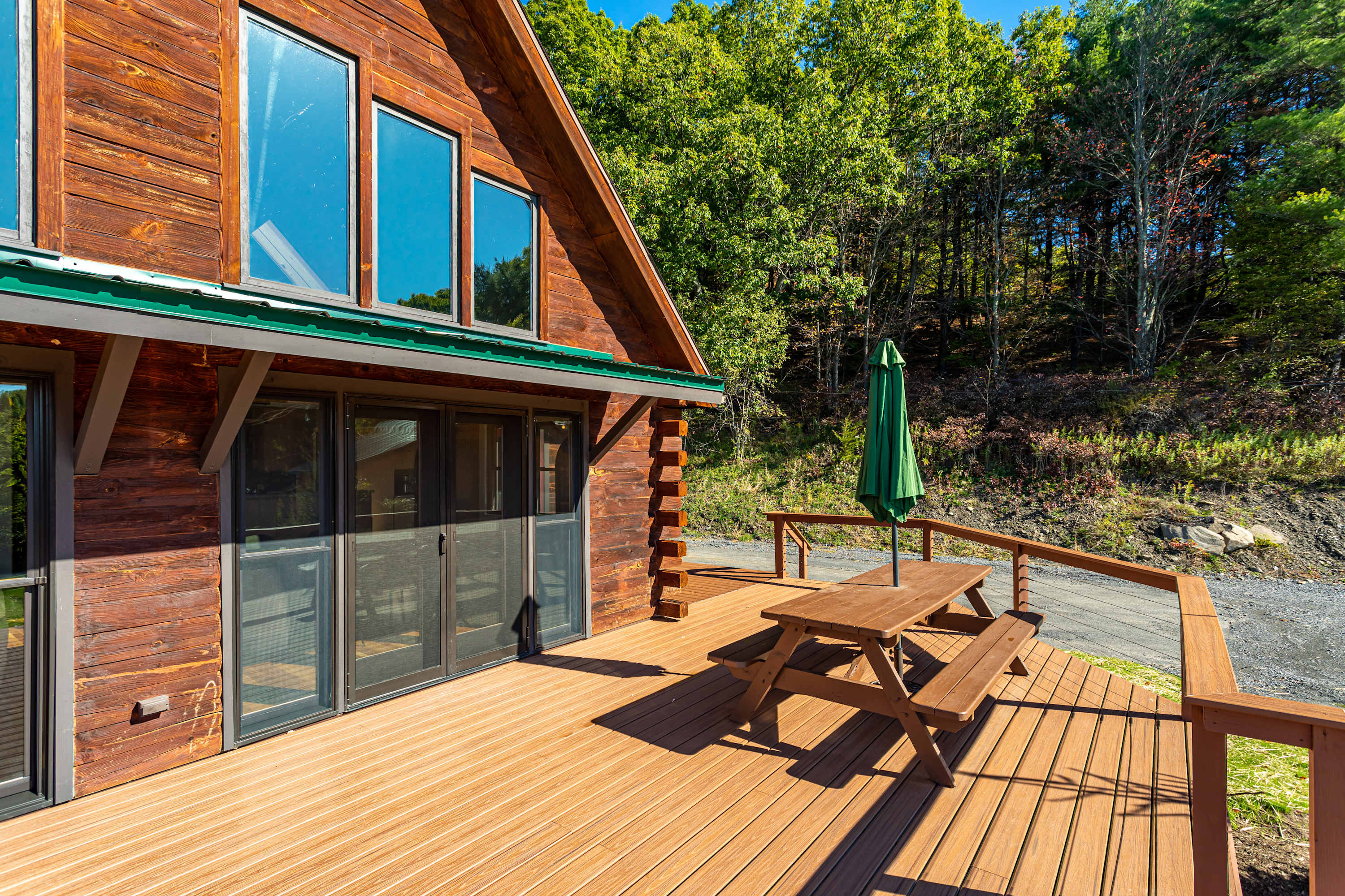 Expansive wooden deck with a picnic table and green umbrella, overlooking a wooded hillside. The log cabin exterior features large windows and glass doors.