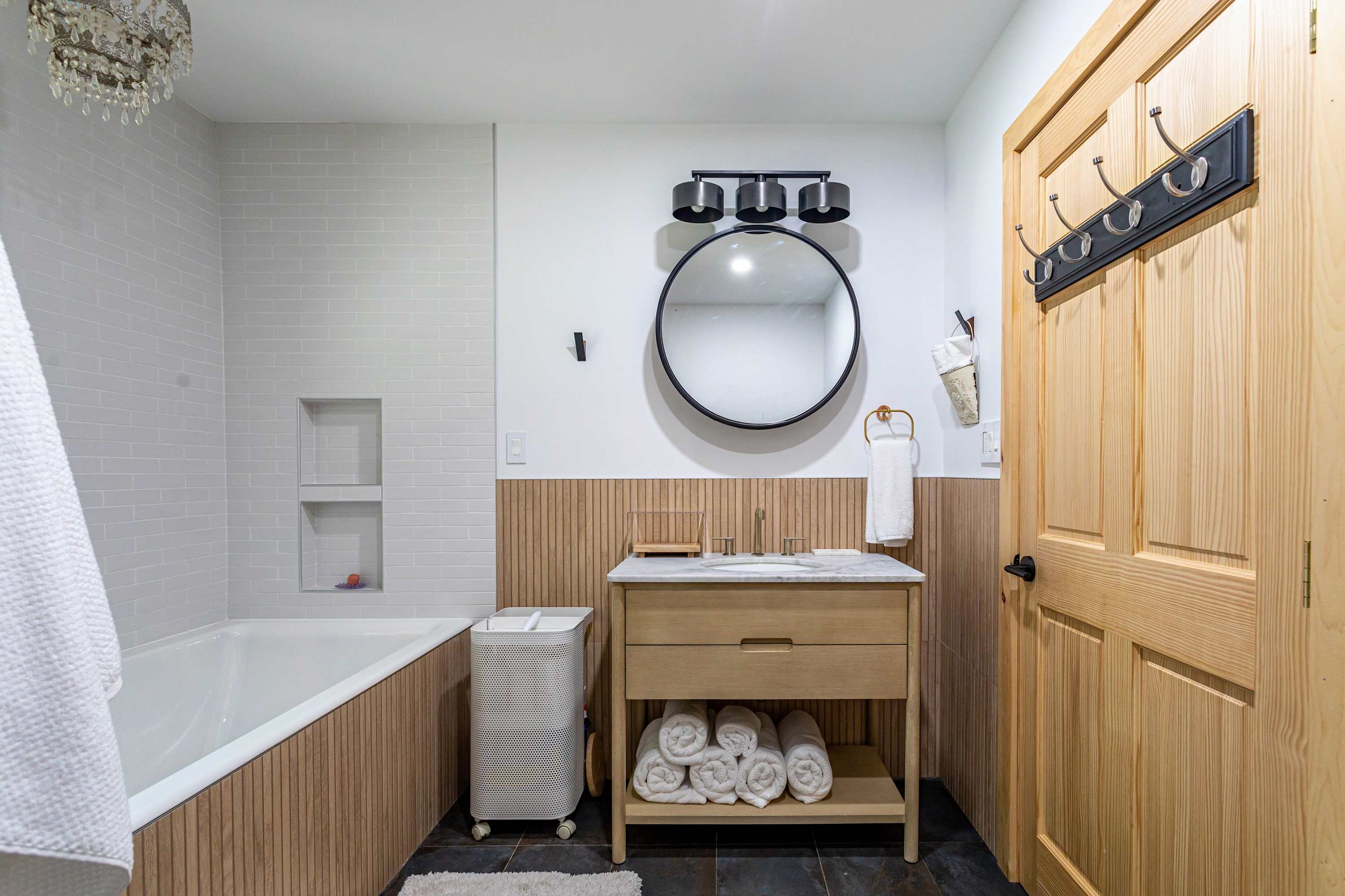 View of bathroom-2's vanity with a marble countertop, wooden base, and a round black-framed mirror. The shower/tub combo with subway tiles and wooden paneling is visible to the left.