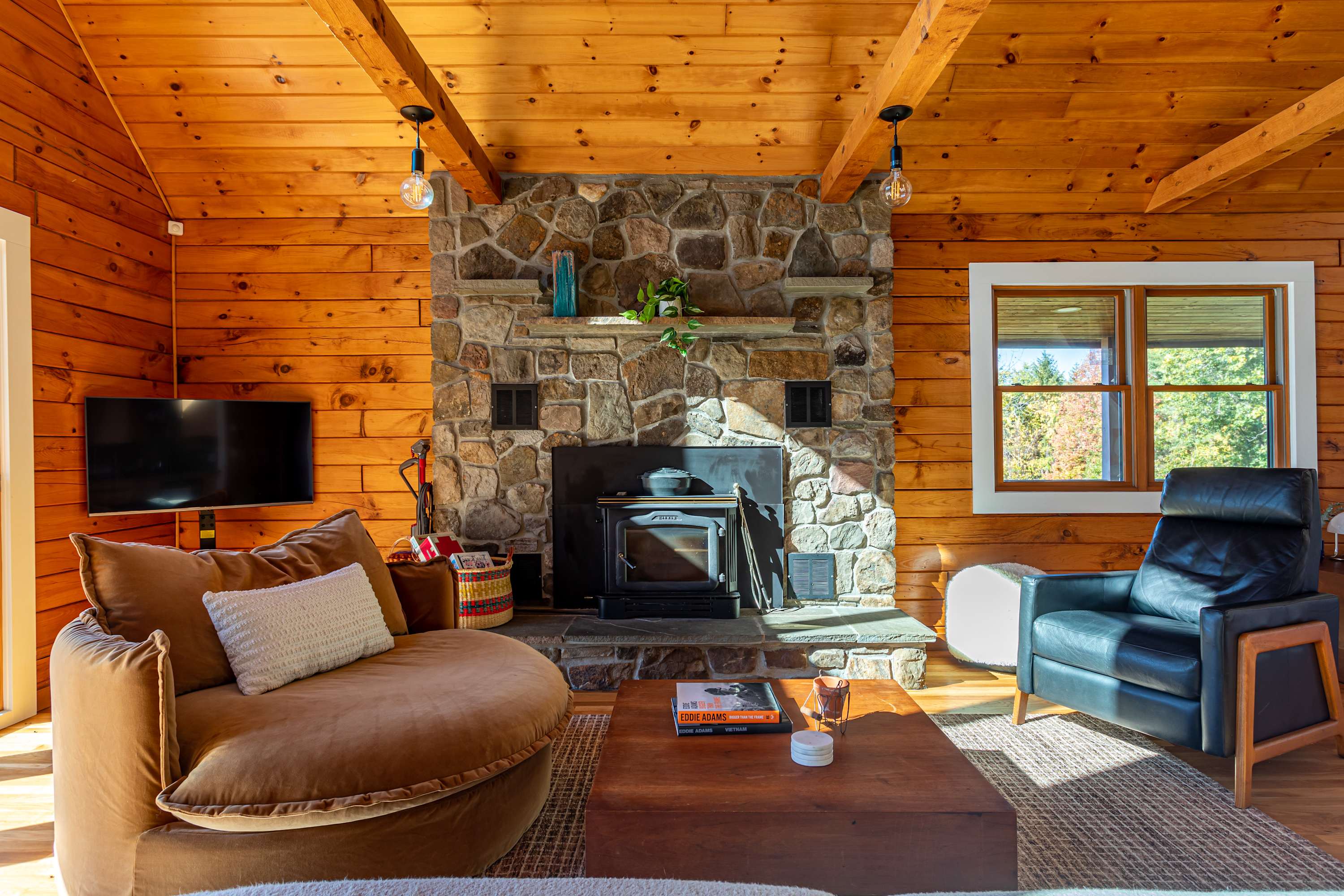Inviting living room featuring a prominent stone fireplace with a wood-burning stove insert, a comfortable round velvet armchair, and a wooden coffee table, surrounded by warm wood-paneled walls and hardwood floors.