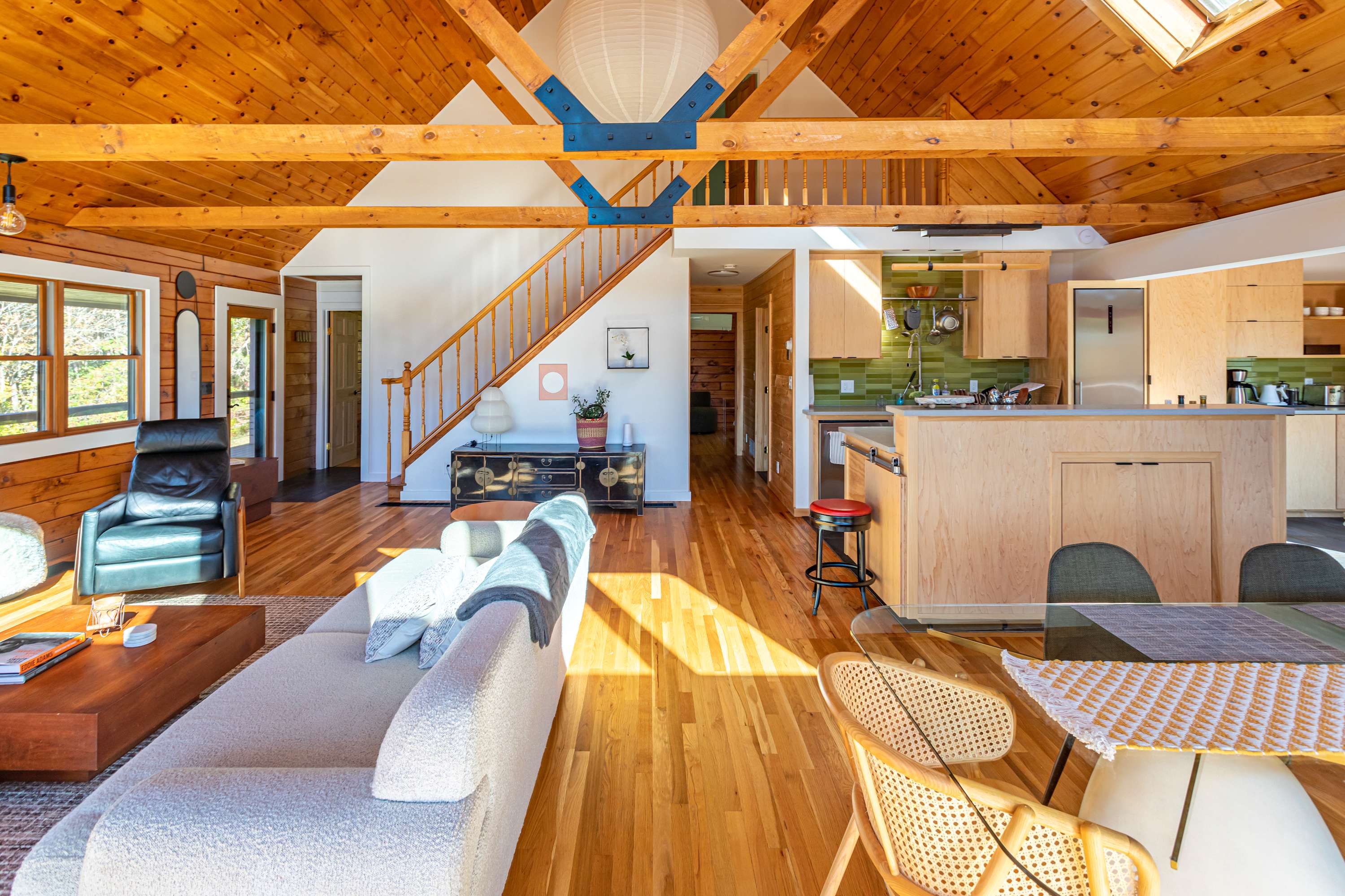 Bright open-concept living area with light hardwood floors, a white sectional sofa, and a view towards the kitchen island and a wooden staircase leading to a loft, all beneath a high vaulted wooden ceiling.