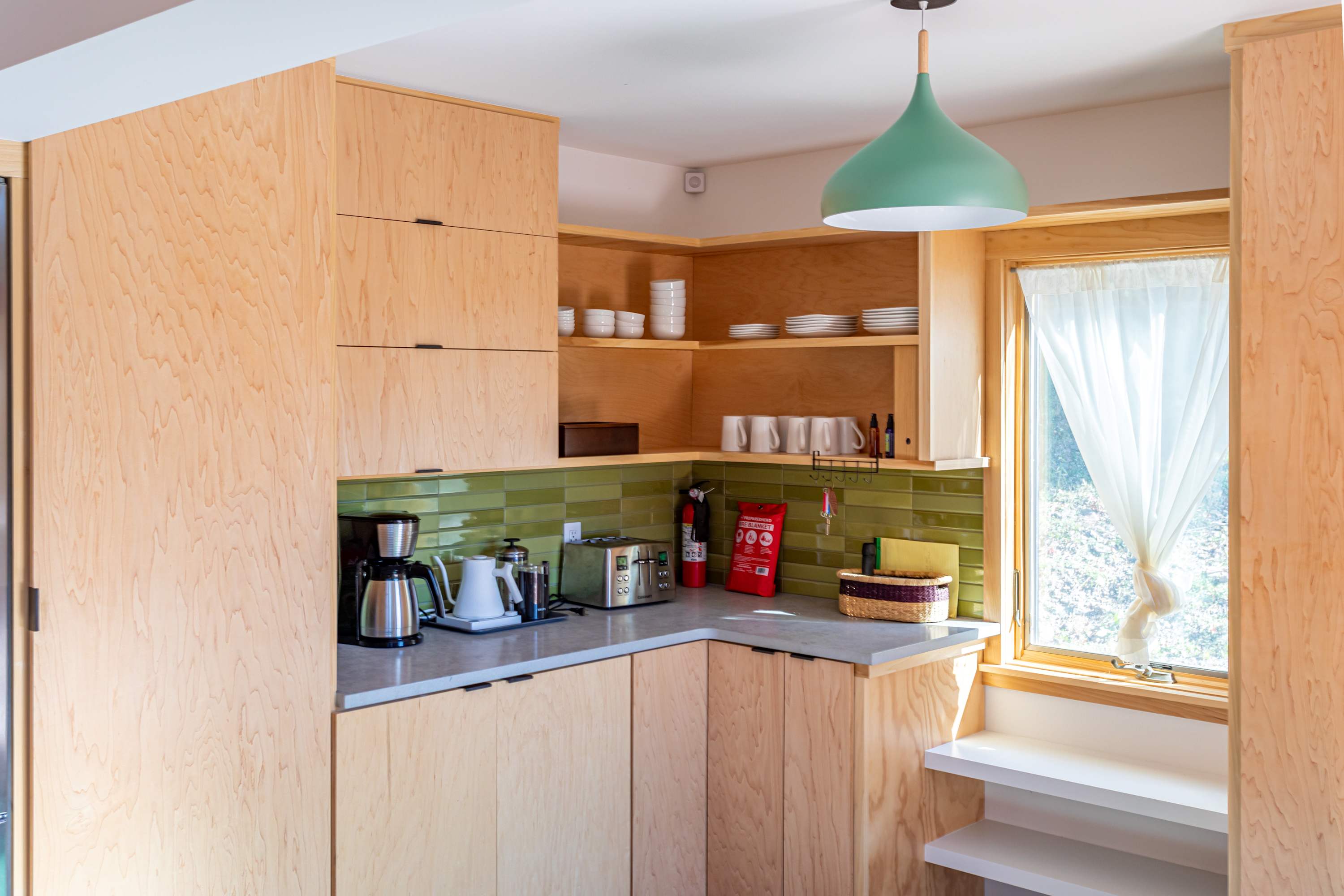 Cozy kitchen corner with light wood open shelving and closed cabinets, a light gray countertop, and a green subway tile backsplash, featuring a coffee maker, toaster, and a window with white curtains.