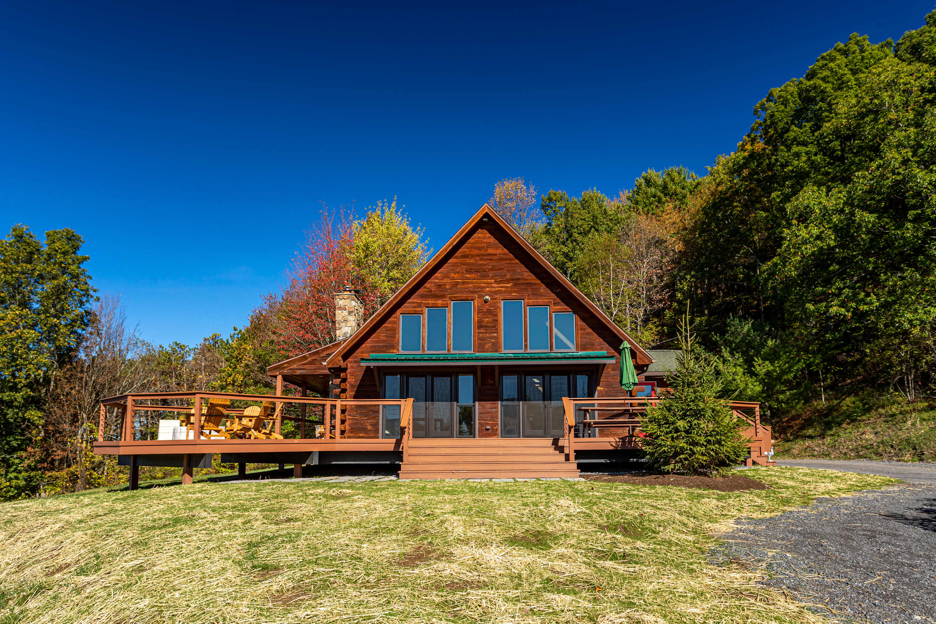 Front exterior of a rustic wooden A-frame cabin with a green roof, large windows, and a spacious wraparound wooden deck, surrounded by autumn trees and a clear blue sky.