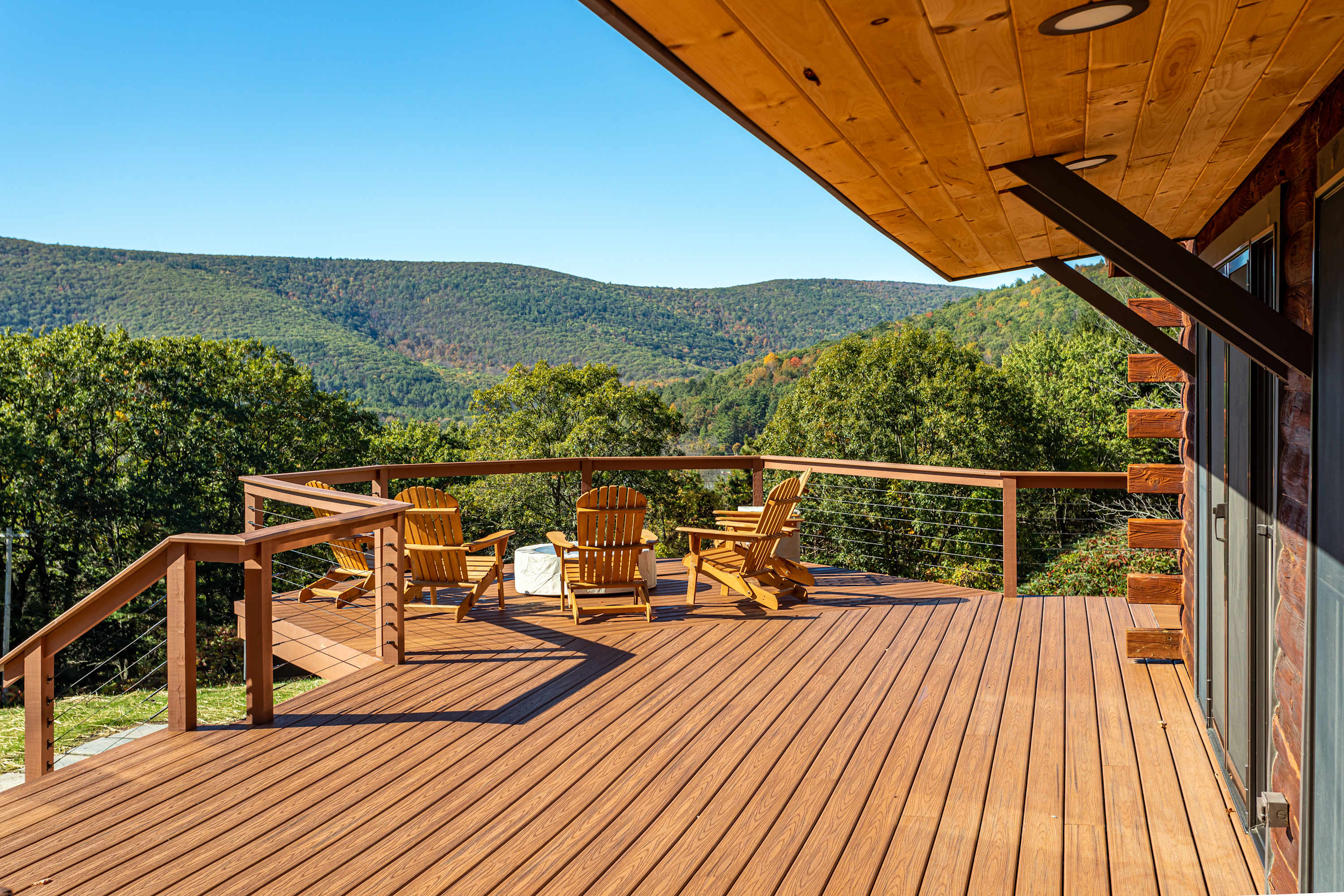 Expansive wooden deck with cable railings, furnished with several Adirondack chairs, offering panoramic views of rolling green mountains and a clear blue sky.