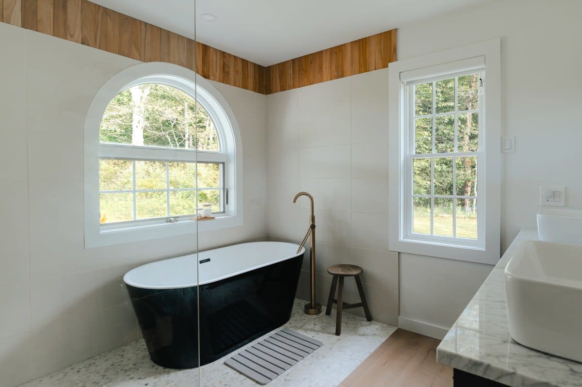 Close-up of a luxurious bathroom with a black freestanding tub, a brass floor-mounted faucet, and a large arched window offering natural light. Light wood paneling accents the wall above the white tiled shower area.