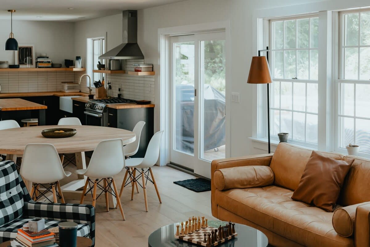 An open-concept living space combines a modern kitchen and dining area with a cozy living room featuring a tan leather sofa and a black and white plaid armchair. Large windows overlook the outdoors, and light hardwood floors connect the spaces.
