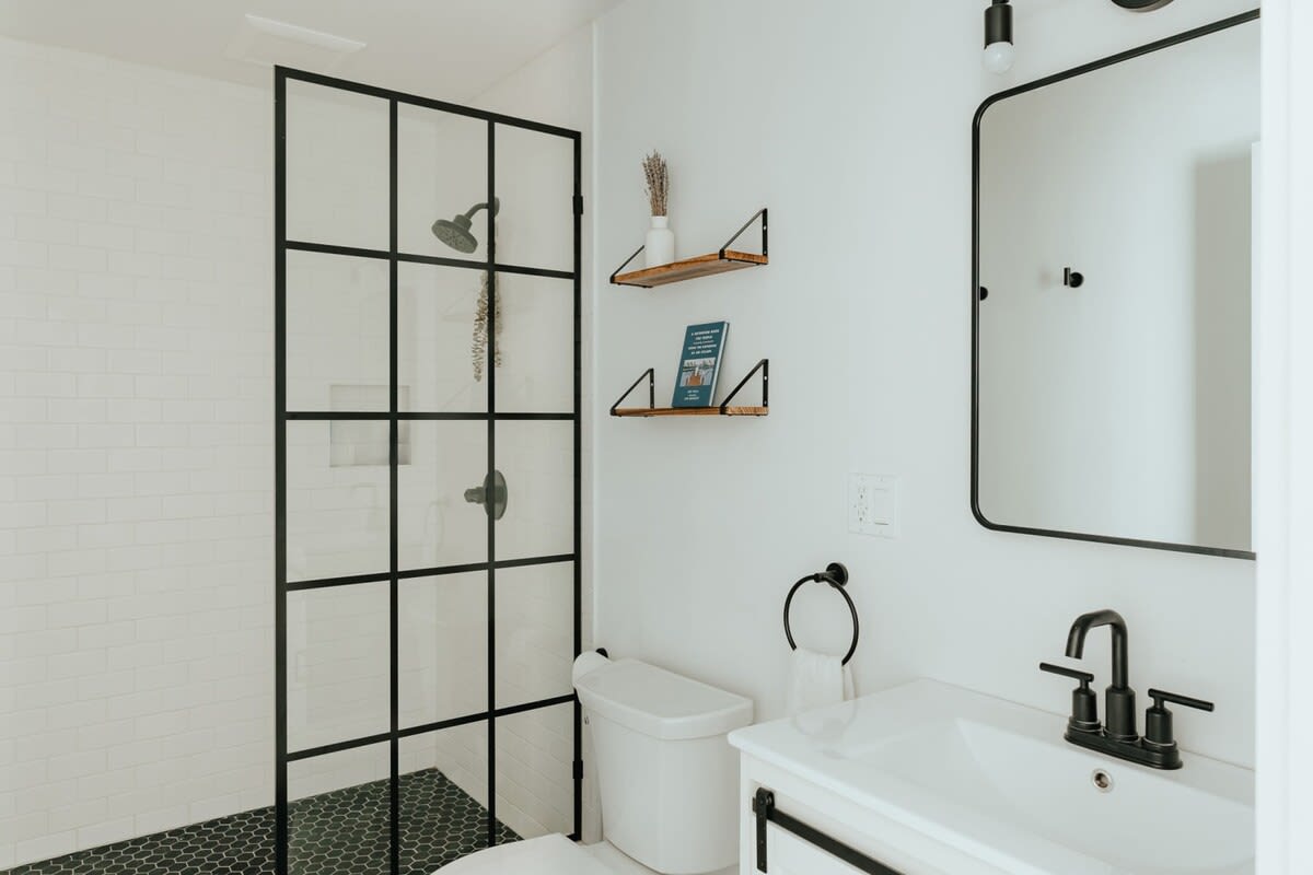 Modern bathroom with a black-framed glass shower, white subway tile walls, and black hexagonal floor tiles. A white vanity with a black faucet and a large rectangular mirror completes the space.