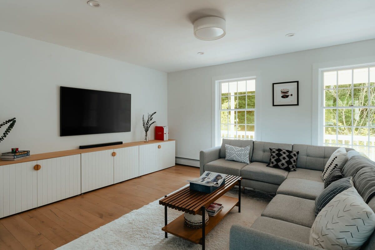 Modern living room featuring a large wall-mounted TV above a long white cabinet with wooden accents, a comfortable grey sectional sofa with patterned pillows, and a wooden coffee table on a fluffy white rug.
