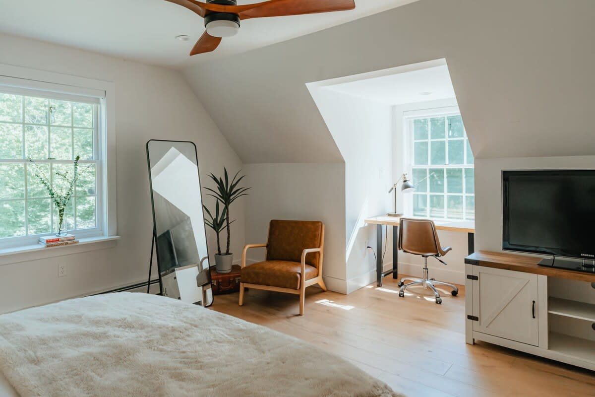 Spacious bedroom featuring a bed with a fluffy blanket, light wood floors, a ceiling fan, and a window. A dedicated workspace with a desk and chair is visible, along with a full-length mirror and a plant.