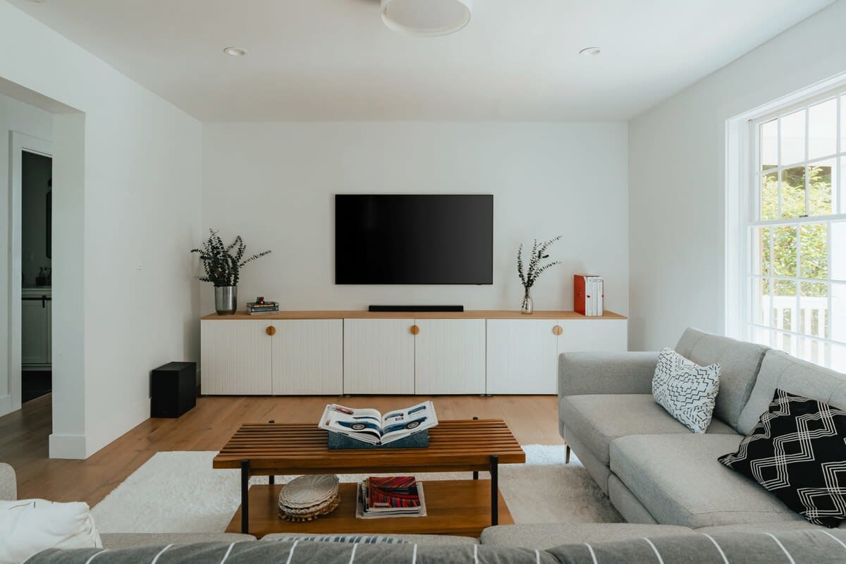 A contemporary living room features a wall-mounted television above a long white media console with reeded cabinet doors. A light gray sofa with patterned pillows and a wooden slatted coffee table are placed on a plush white rug, with large windows providing natural light.