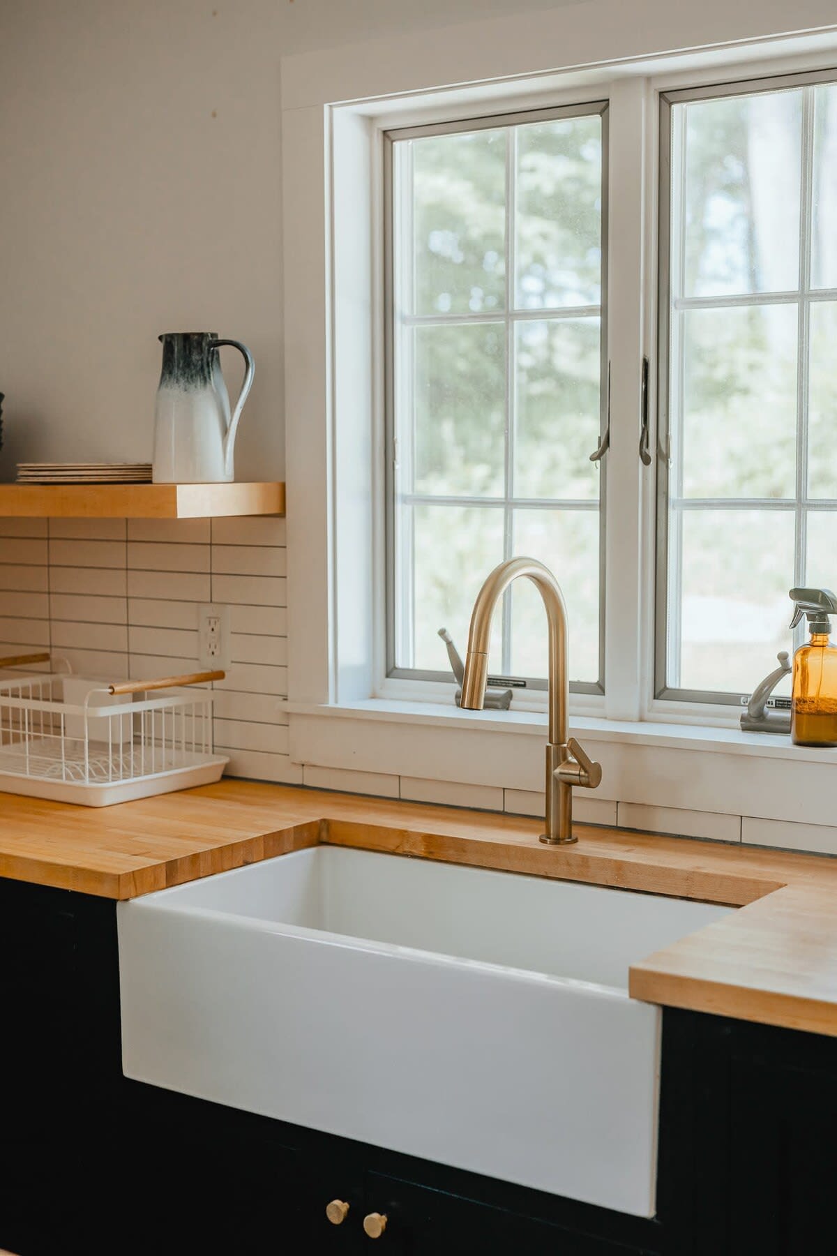 A bright kitchen sink area features a large white farmhouse sink with a modern gold faucet, set against light wood countertops and white subway tile backsplash. A large window offers a view of lush greenery outside.