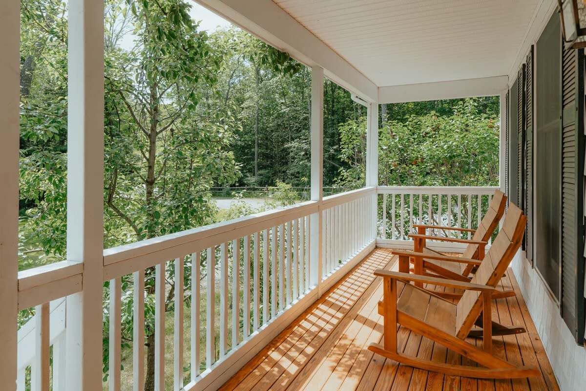 Inviting covered porch with light wooden floors and white railings, furnished with two classic wooden rocking chairs, offering a serene view of the surrounding dense green trees.
