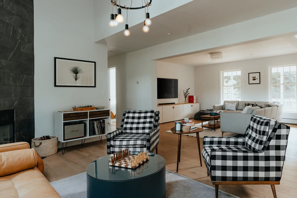 A stylish living room features a dark stone fireplace, two black and white plaid armchairs, and a tan leather sofa, arranged around a round coffee table with a chess set. A unique chandelier hangs from the ceiling, and light hardwood floors complete the space.