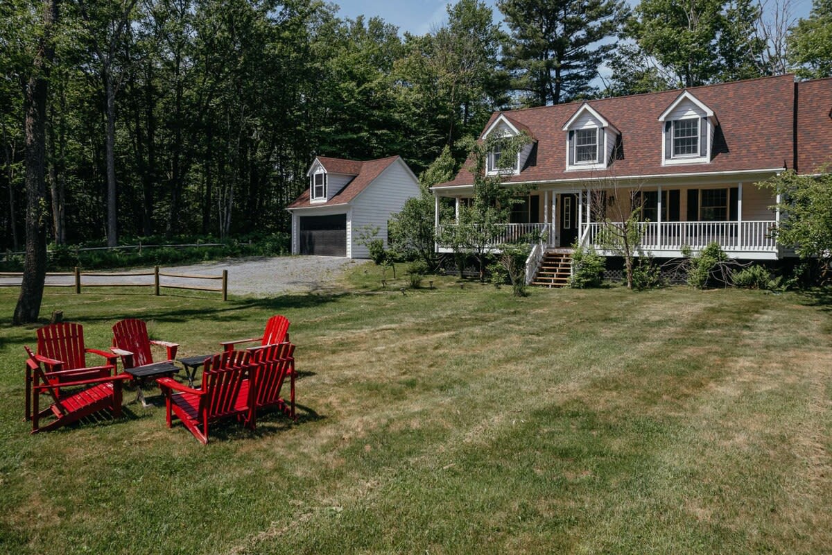 Expansive front view of a white-sided house with a brown roof and dormer windows, featuring a wide porch with white railings, a detached garage, and a large grassy yard with a cluster of red Adirondack chairs.