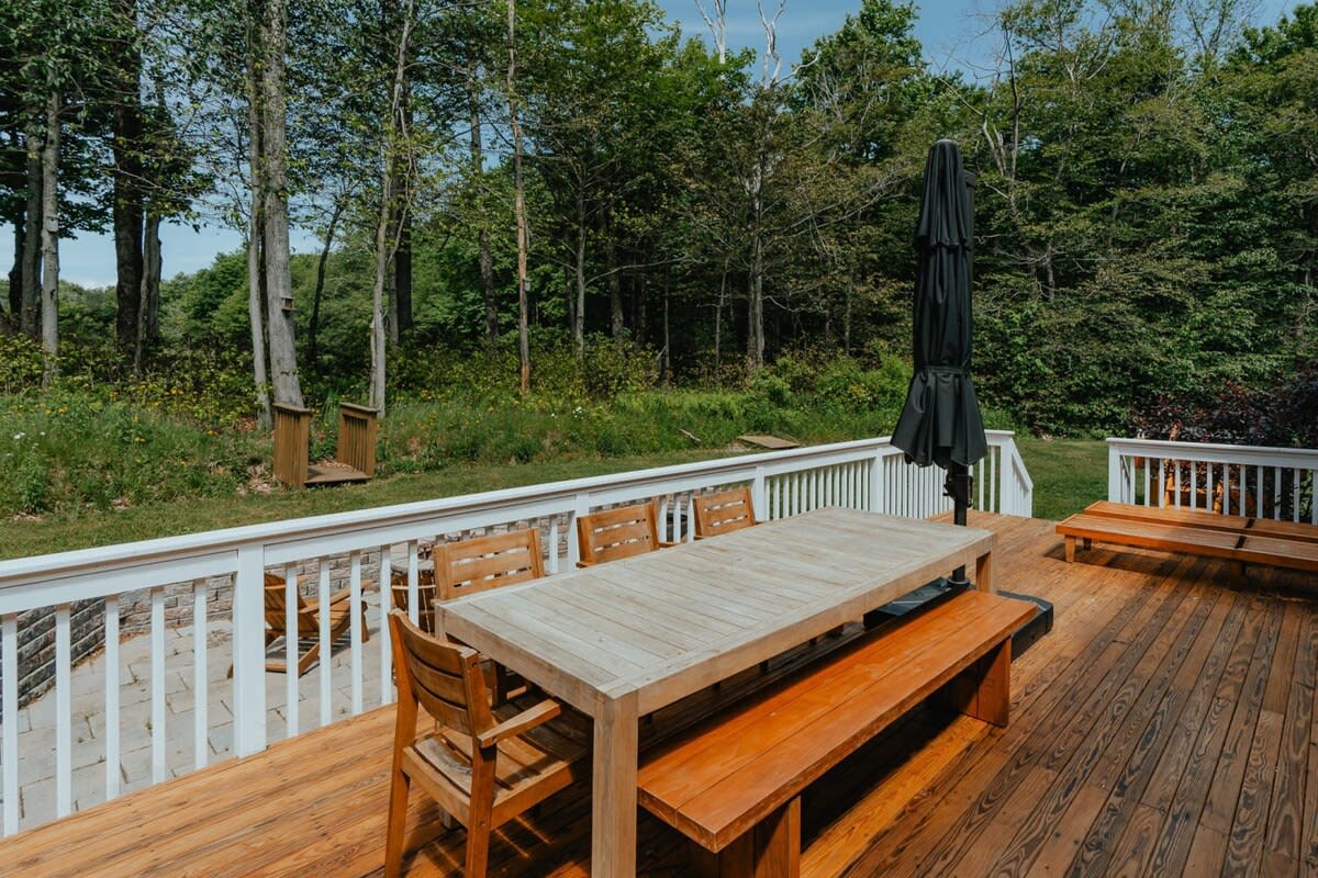 Spacious outdoor deck with a large wooden dining table, benches, and chairs, perfect for al fresco meals. A black umbrella provides shade, and the deck overlooks a lush green backyard and forest.