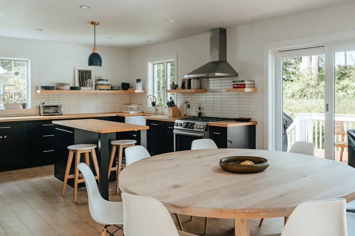 A spacious, open-concept kitchen features black cabinetry, light wood countertops, and white subway tile backsplash, centered around a large round dining table with white chairs. A kitchen island with bar stools and a stainless steel range hood are also visible.