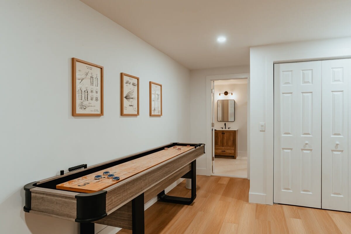 Game room showcasing a long shuffleboard table with pucks, set against white walls with framed patent art. A doorway leads to a bathroom with a wooden vanity and a mirror.