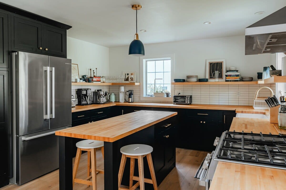 A modern kitchen features sleek black cabinetry, light wood countertops, and white subway tile backsplash, with a stainless steel refrigerator and a kitchen island with two bar stools. Open shelving displays dishes and decor, illuminated by a blue pendant light.