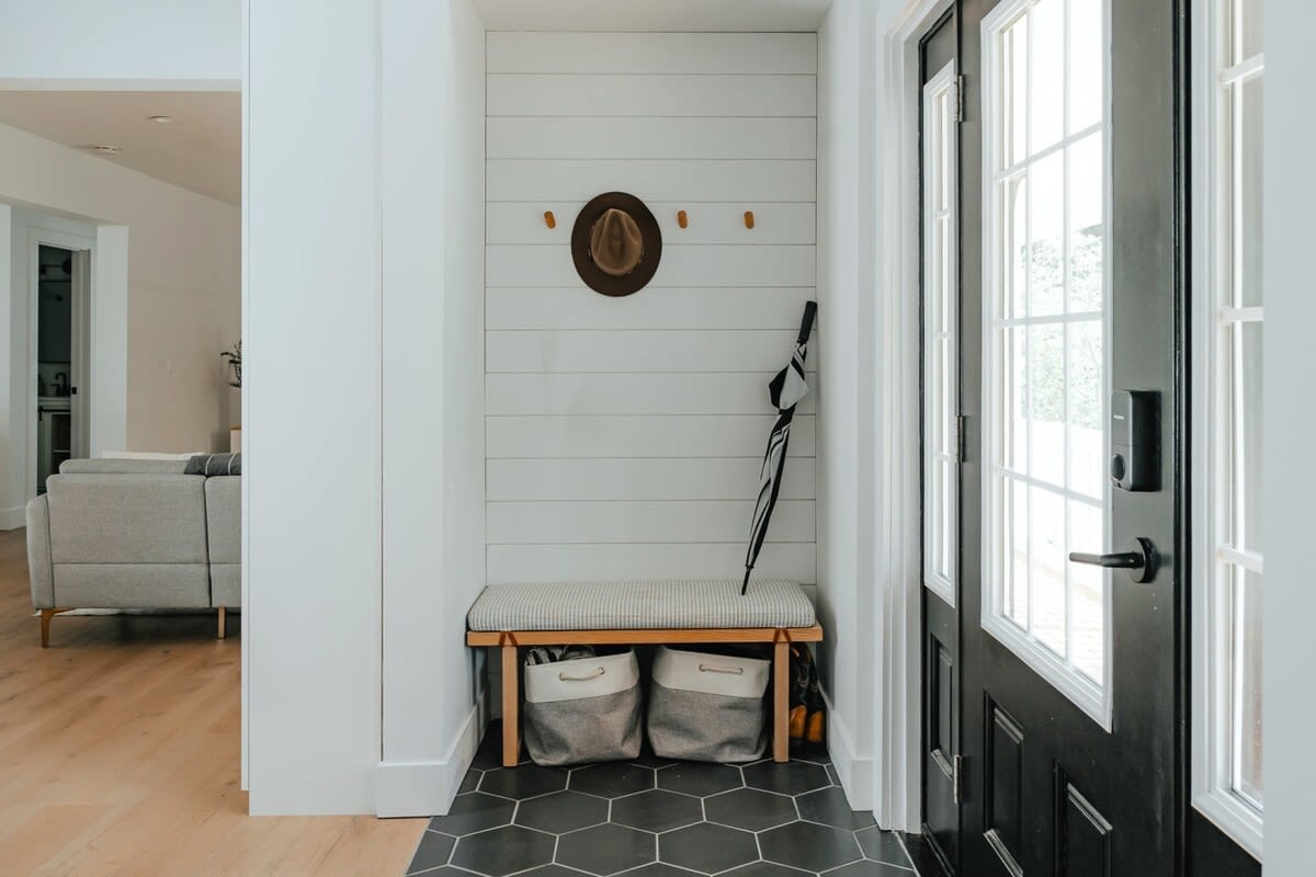 A bright entryway features white shiplap walls with wooden hooks, a bench with a striped cushion, and two storage baskets underneath, all set on dark hexagonal floor tiles. A black front door with glass panels is visible on the right.