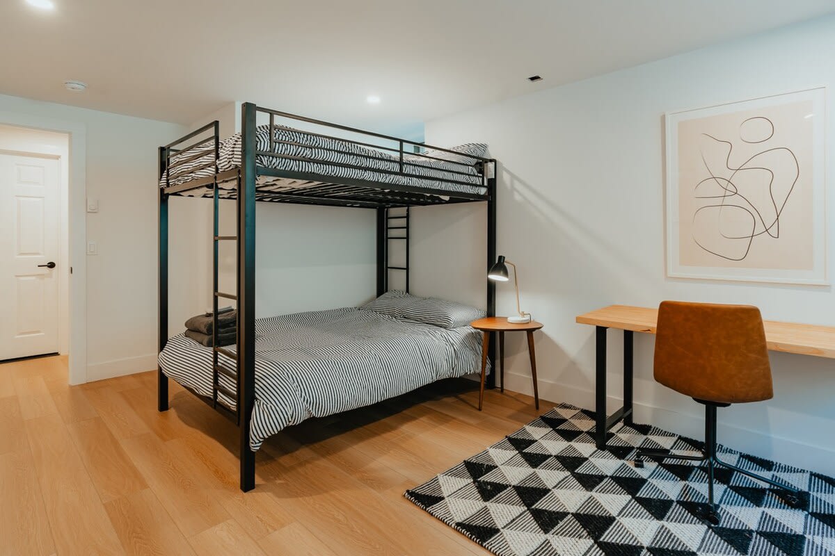 Kids' bedroom featuring a black metal bunk bed with striped bedding and light wood floors. A desk with a chair and a small side table with a lamp provide a functional workspace.