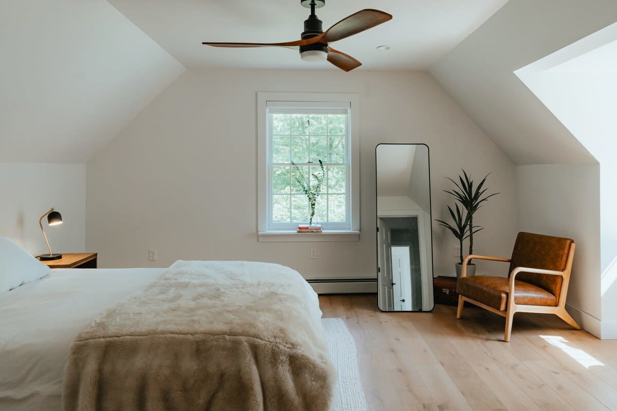 Bright bedroom with white walls, light wood floors, a bed with a fluffy blanket, a ceiling fan, and a large window overlooking trees. A full-length mirror and a brown leather armchair add to the decor.