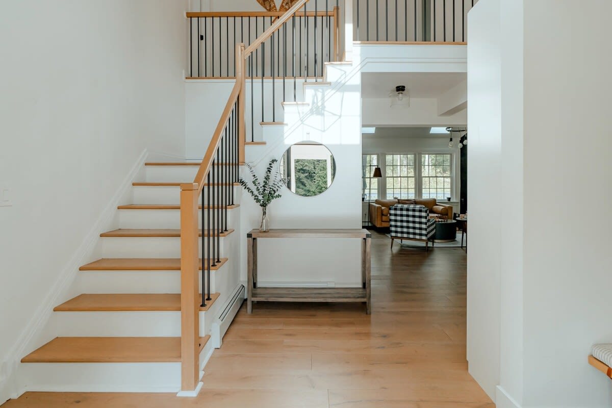 A grand entryway features a light wood staircase with black metal balusters, leading to an upper floor. A round mirror hangs above a rustic console table, with light hardwood floors extending into an open-concept living space.