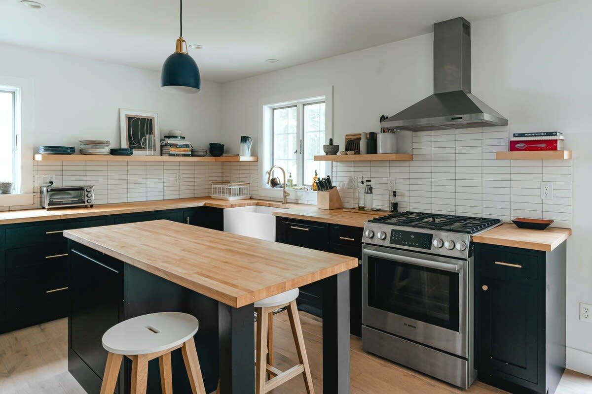 A well-appointed kitchen showcases black base cabinets, light wood countertops, and white subway tile backsplash, with a stainless steel gas range and range hood. A kitchen island with bar seating extends into the space, all on light hardwood floors.