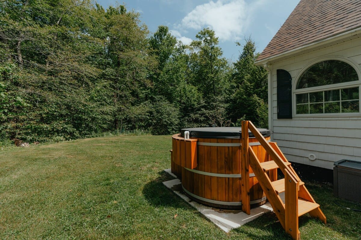 A rustic cedar hot tub with a dark cover and wooden steps sits on a concrete pad in a vibrant green lawn, backed by a white-sided house and dense green forest under a blue sky.