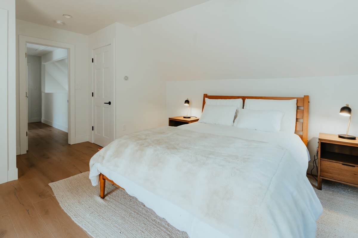 Cozy bedroom with a wooden bed frame, white bedding, and two dark wood nightstands with black lamps. Light wood floors and a woven rug complete the simple, clean aesthetic.