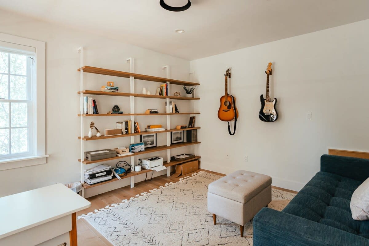 Bright office space with a large, multi-tiered wooden bookshelf filled with books and decor. Two guitars are mounted on the wall, and a blue sofa with an ottoman provides seating.