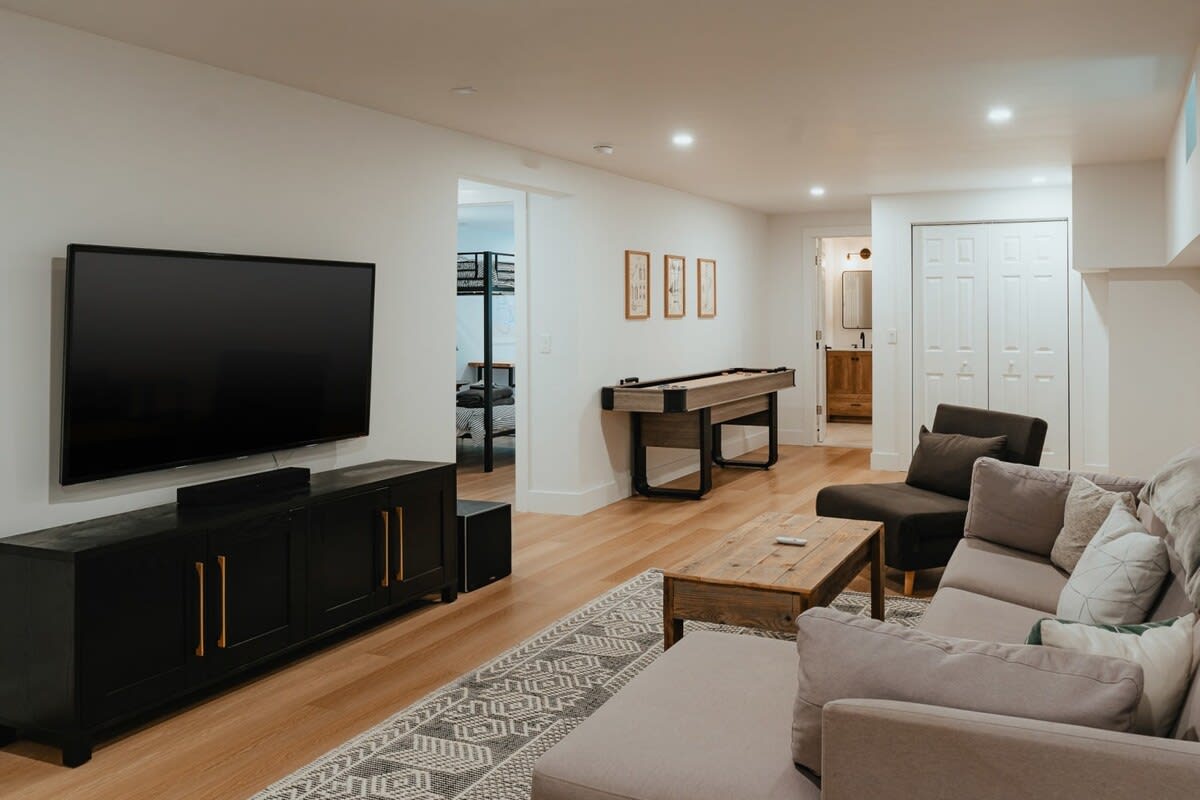 Modern game room featuring a large flat-screen TV on a black console, a light gray sectional sofa, and a rustic wooden coffee table. A shuffleboard table is visible in the background, leading to another room.