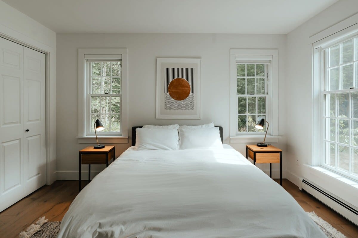 Bright bedroom with a queen bed, white bedding, and two wooden nightstands with black lamps. An abstract art piece hangs above the bed, flanked by two large windows overlooking greenery.