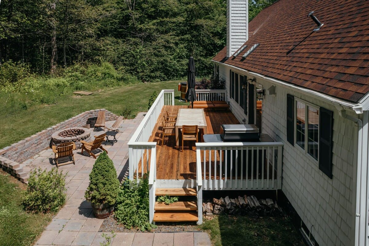 Elevated view of a spacious wooden deck with white railings, featuring a large outdoor dining table and chairs, a grill, and steps leading down to a stone patio with a fire pit, all against a backdrop of lush trees.