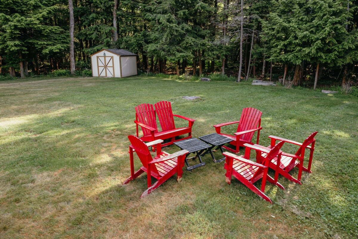 Expansive grassy yard featuring a circular arrangement of bright red Adirondack chairs around a small black table, ideal for outdoor relaxation. A shed is visible in the background, surrounded by tall trees.