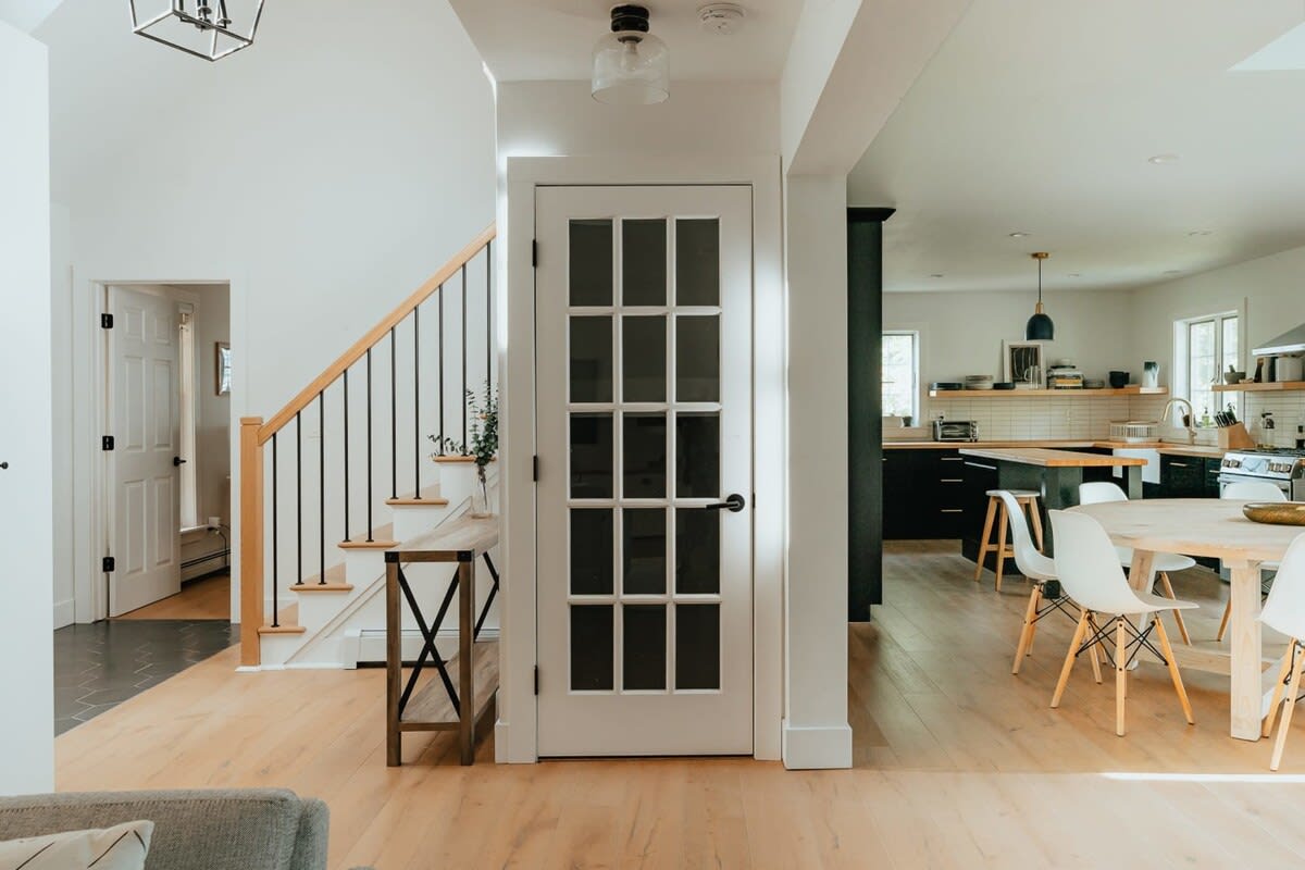A bright hallway showcases a light wood staircase with black balusters and a white French door with multiple glass panes. Beyond, an open-concept layout reveals a modern kitchen and dining area with light hardwood floors.