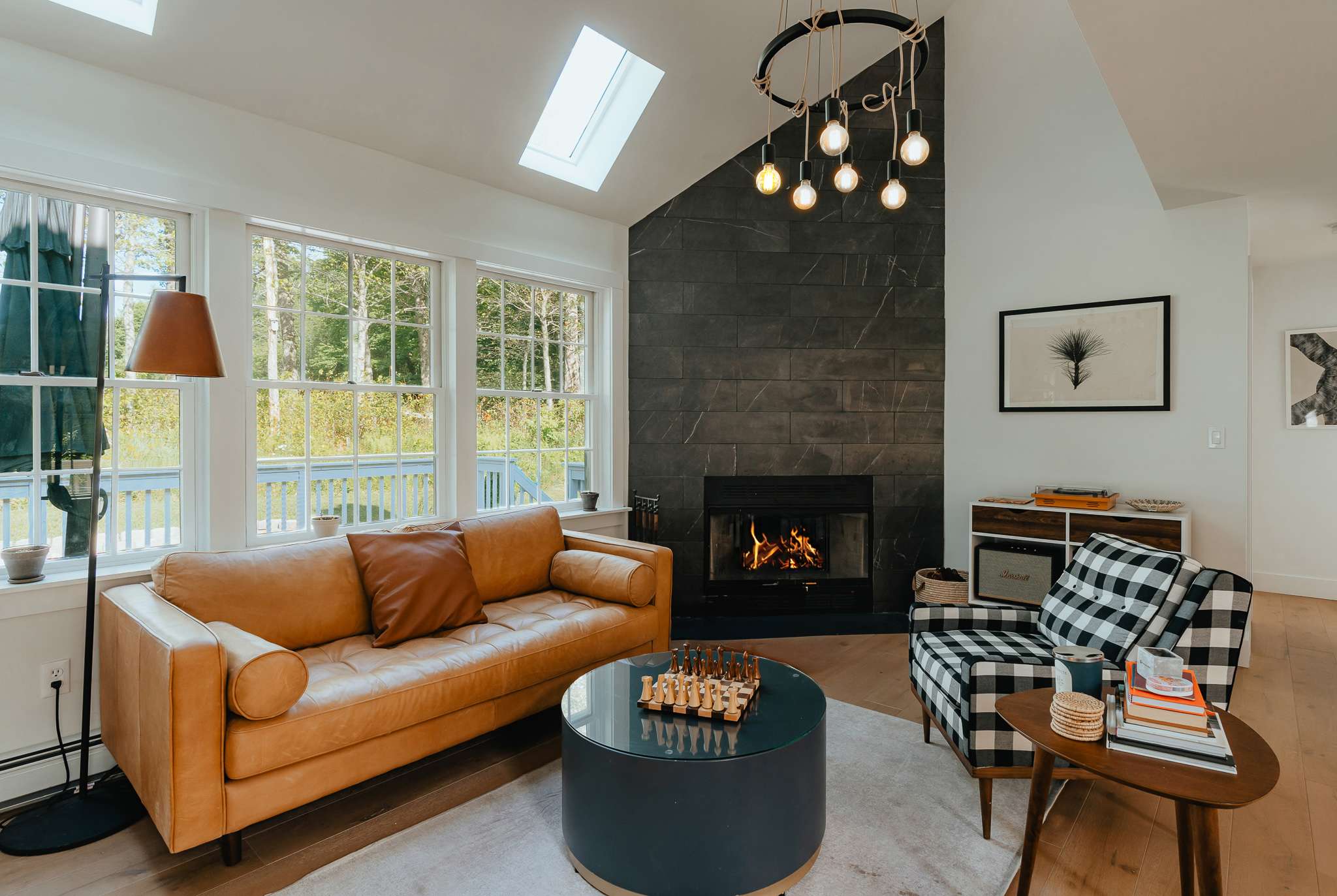 Bright living room with a tan leather sofa, a black and white plaid armchair, and a dark round coffee table with a chess set, centered around a modern black tiled fireplace with a unique circular chandelier.