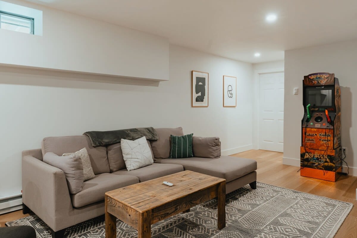 Inviting game room with a light gray sectional sofa, a rustic wooden coffee table, and an arcade machine. The space features light wood floors, a patterned area rug, and framed artwork on the white walls.