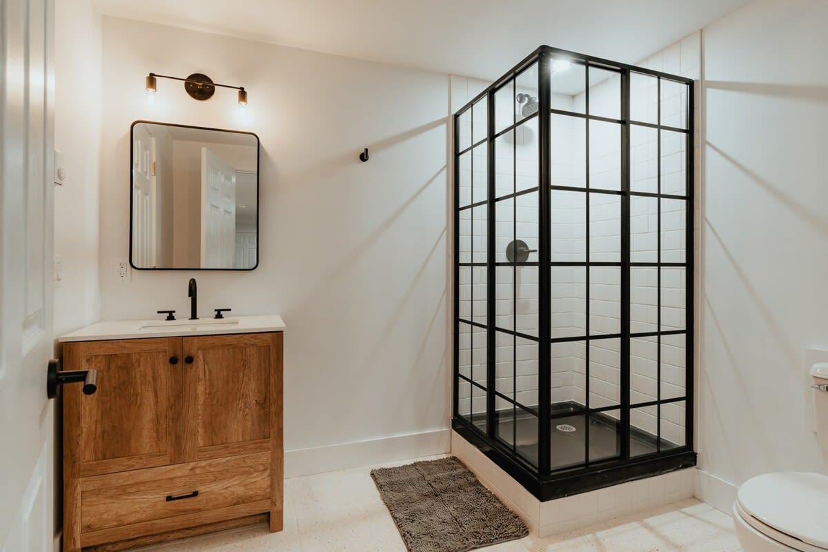 Modern bathroom with a wooden vanity, black faucet, and a rectangular mirror with industrial-style lighting. A black-framed glass shower enclosure with white subway tiles is a prominent feature.