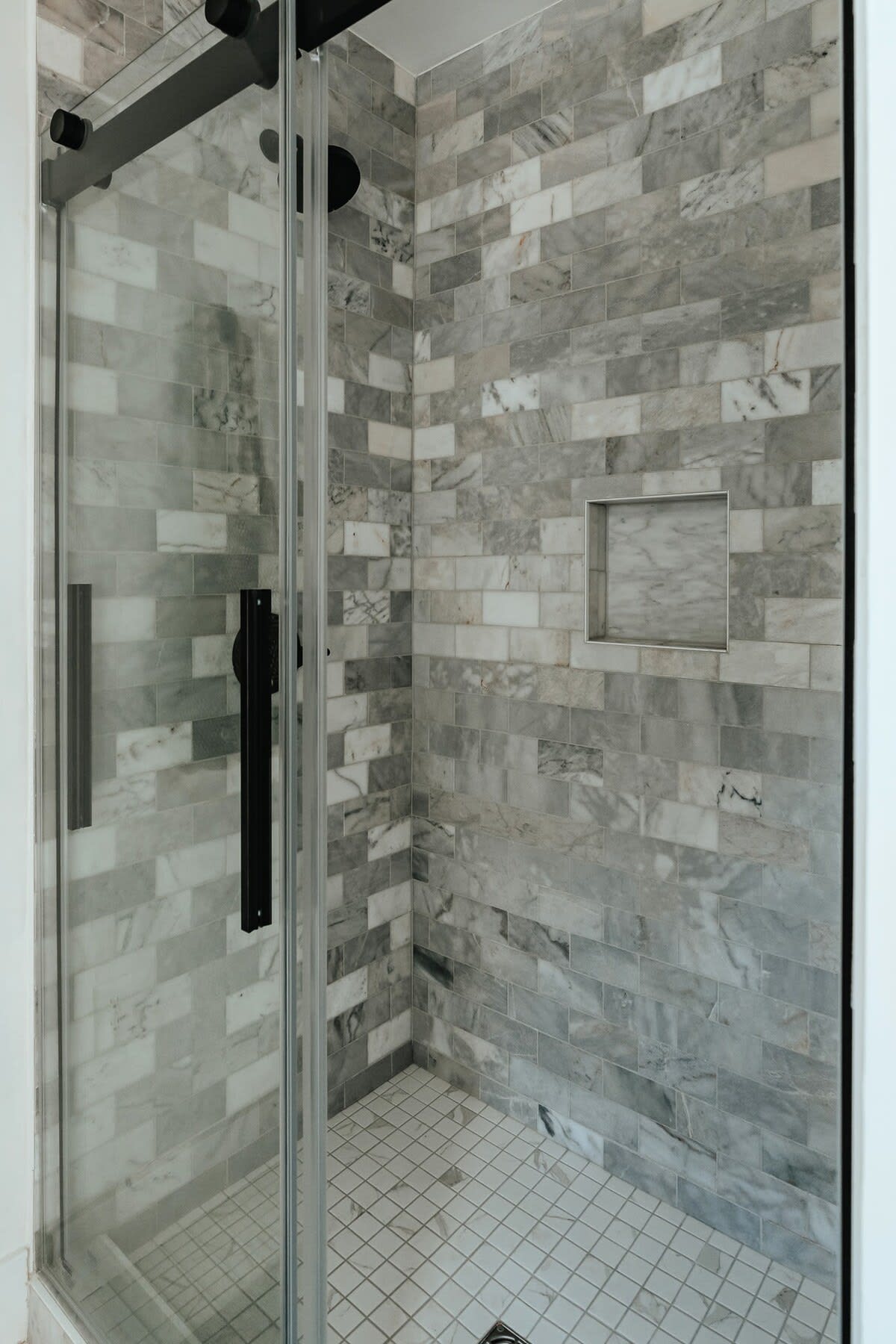 Modern walk-in shower featuring grey and white marble-look subway tiles on the walls and small white square tiles on the floor. A glass enclosure with black hardware is visible.