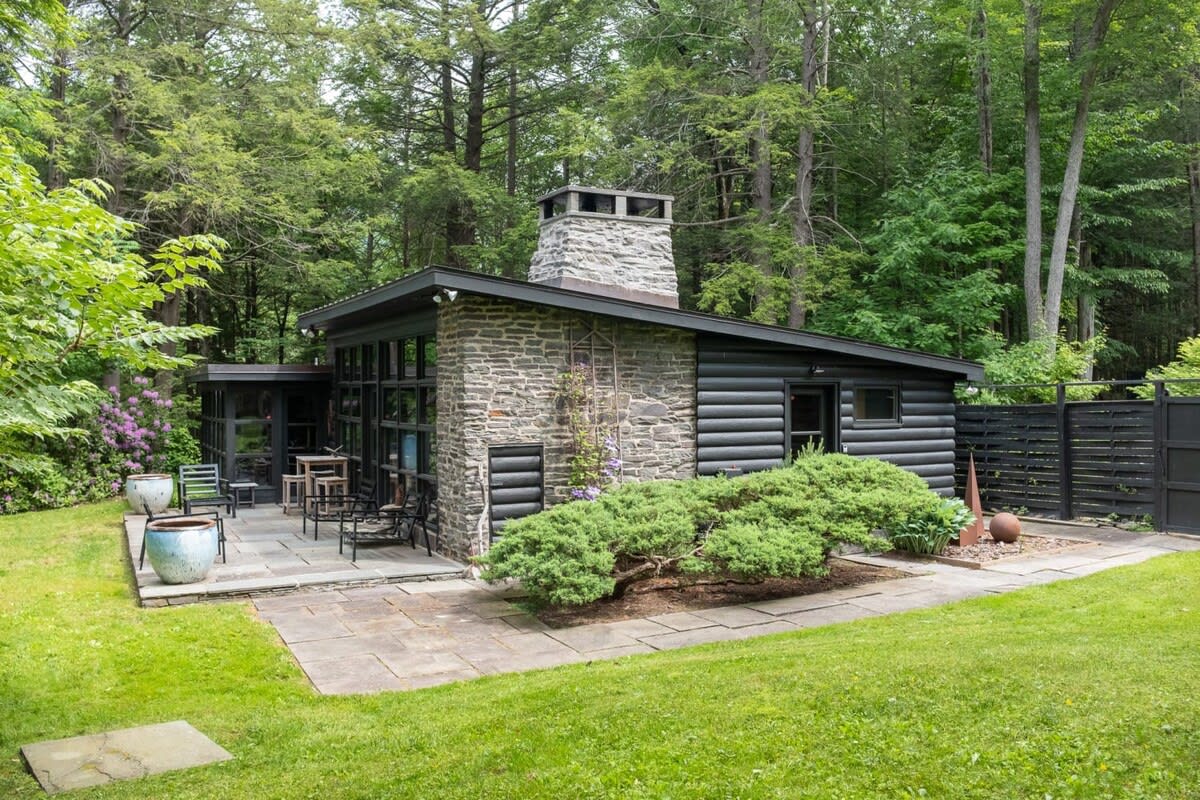 The exterior of a modern cabin with a prominent stone chimney and dark wood siding, nestled among lush green trees. A stone patio with outdoor seating and a well-maintained lawn are visible, offering a serene forest retreat.