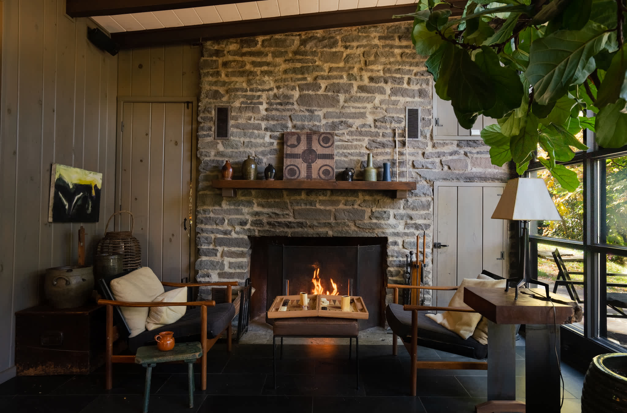 A rustic stone fireplace with a warm fire burning, flanked by two dark armchairs with light cushions. A wooden mantelpiece displays decorative items, and large windows offer a peek at the surrounding greenery, enhancing the cozy ambiance.